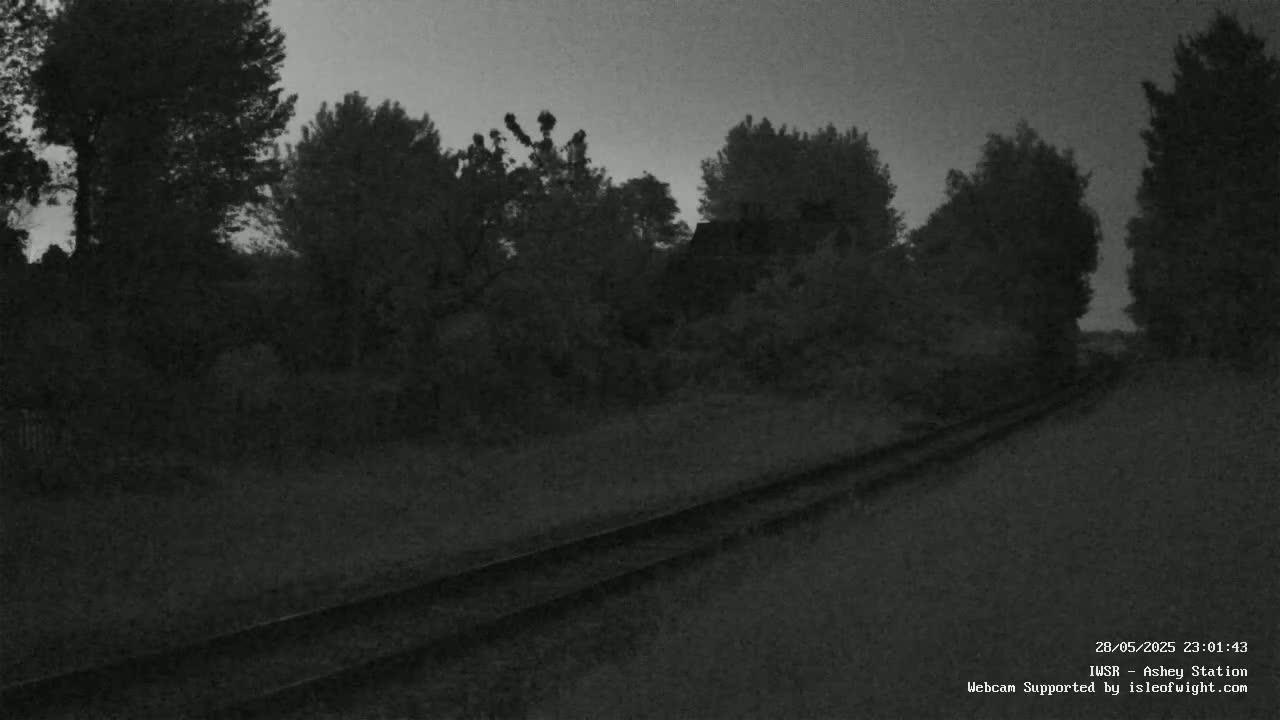 A dark, overcast sky looms over a railway track that winds through a line of silhouetted trees and shrubs.