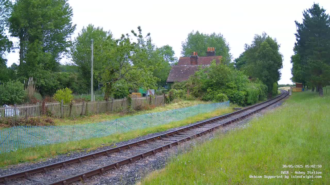 A railway track winds through a grassy field, past a small brick building and wooden fence, under an overcast sky.
