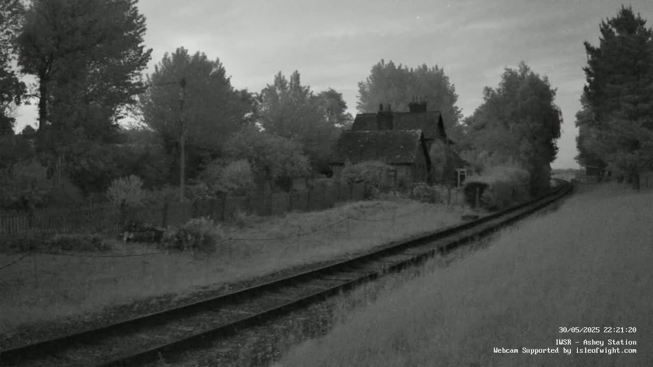 A grayscale image shows a small cottage nestled among trees beside a railway line on an overcast evening.