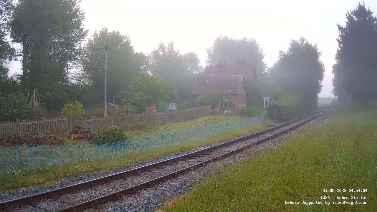 A foggy morning scene shows a small house next to railroad tracks that curve into the distance, surrounded by trees and a wooden fence.