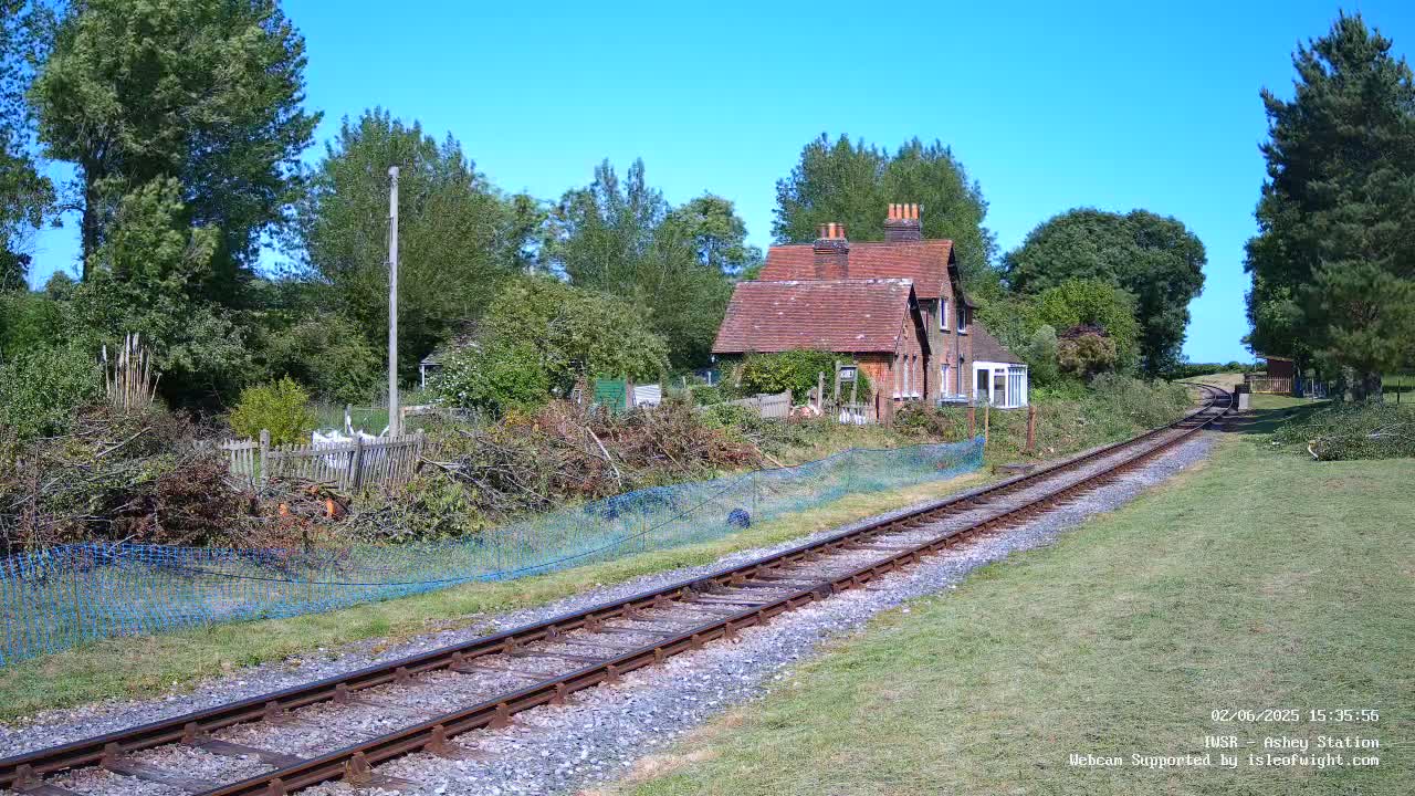 A brick house sits beside a railway line on a sunny day, with overgrown vegetation and a blue mesh fence separating the house from the tracks.