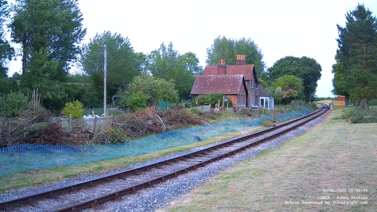 A brick house sits beside a railway track on an overcast day, with a pile of branches behind a blue mesh fence in the foreground.
