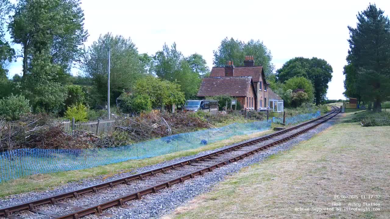 A railway track stretches into the distance, passing a house and a pile of tree branches behind a blue mesh fence, under a partly cloudy sky.