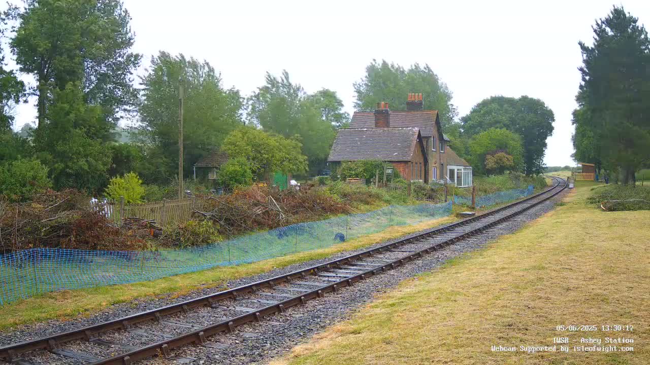 A railway track extends into the distance past a small house and overgrown vegetation under an overcast sky.