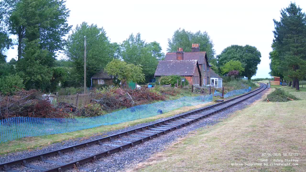 A railway track curves gently through a grassy area, passing alongside a small house and a pile of pruned branches behind a blue mesh fence, under an overcast sky.