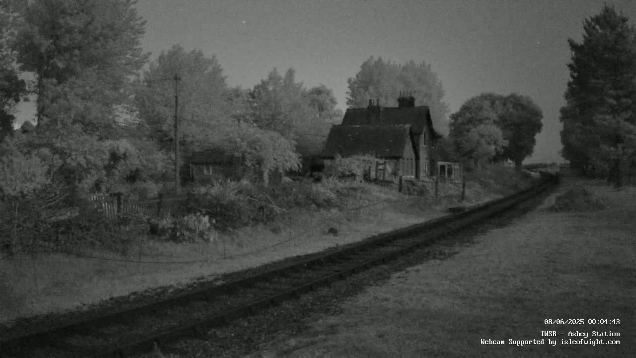 A nighttime, grayscale image shows a small house nestled among trees beside a railway track under a mostly clear sky.