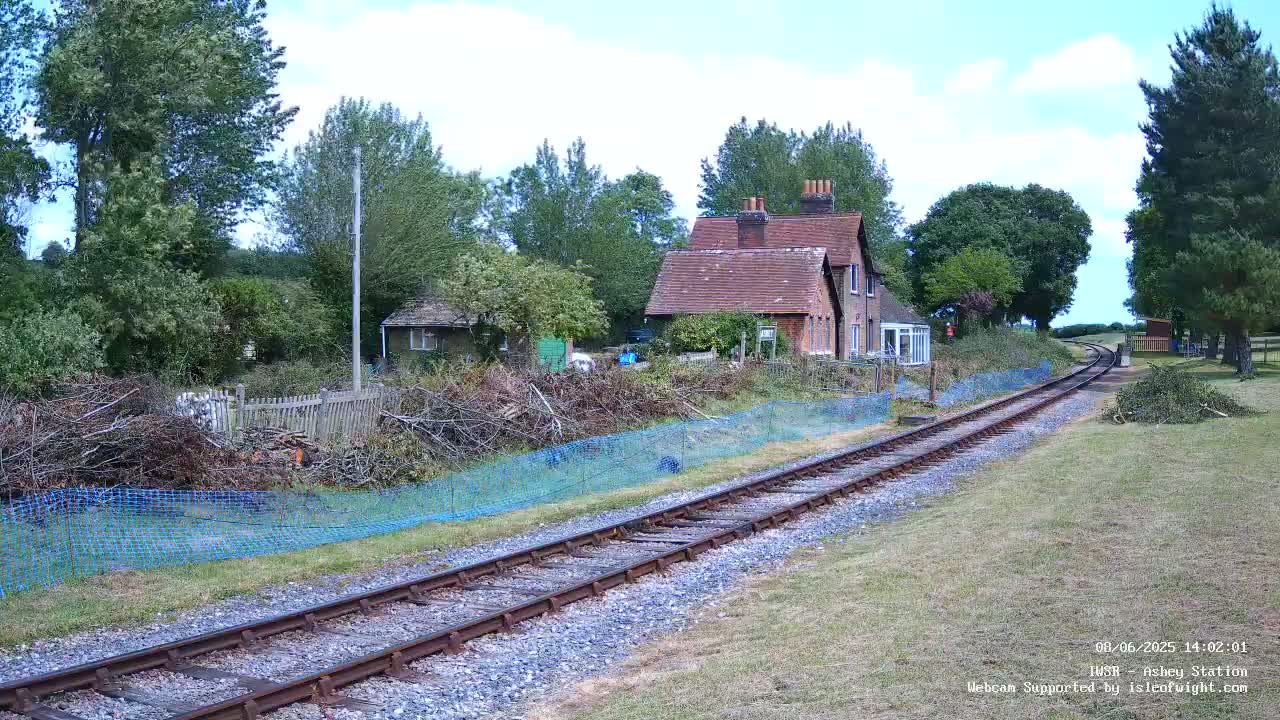 A railway track curves gently past a small brick house and a pile of pruned branches behind a blue mesh fence, under a partly sunny sky.