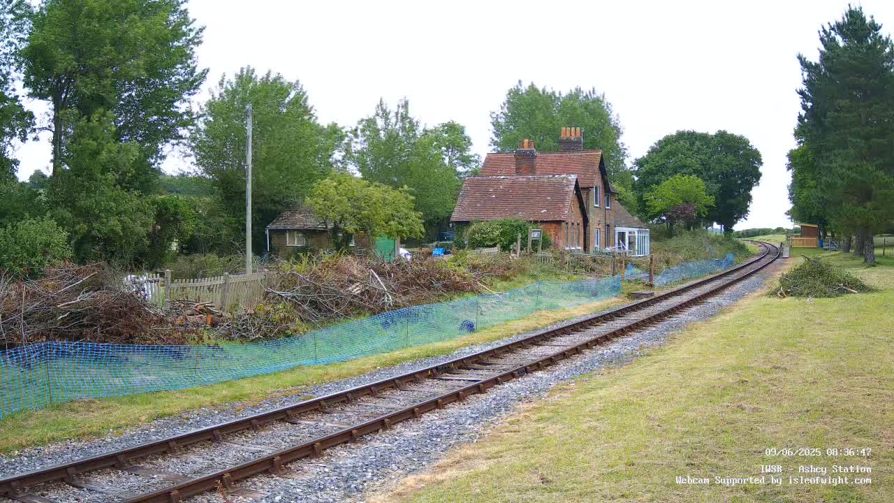 A railway track extends into the distance past a small brick house and a pile of brush behind a blue mesh fence, under an overcast sky.
