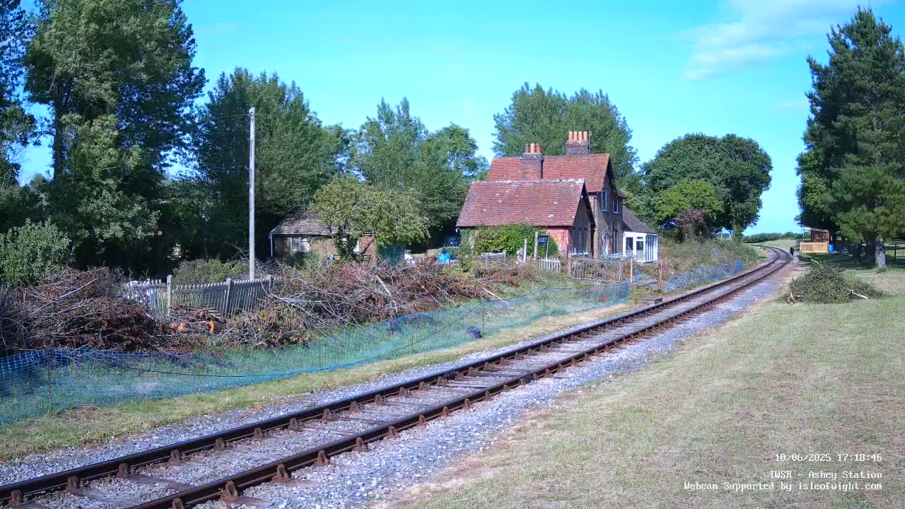 A small brick house sits beside a railway track on a sunny day, with a pile of brush against a fence in the foreground.