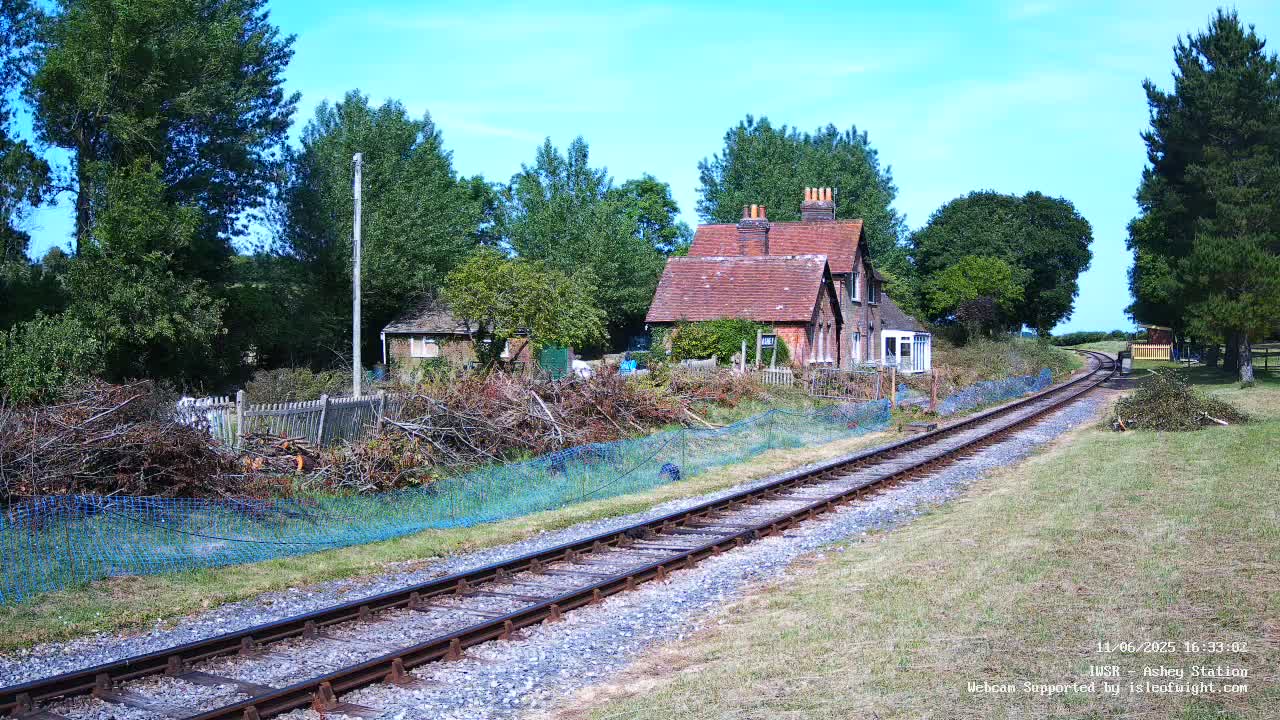 A railway track extends from a brick house into a grassy field under a sunny sky, with piles of pruned branches beside a fence.
