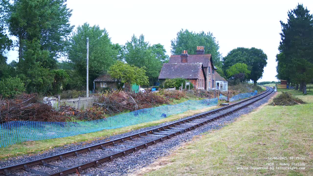 A railway track stretches into the distance, past a brick house and a pile of cut branches behind a blue mesh fence, under an overcast sky.