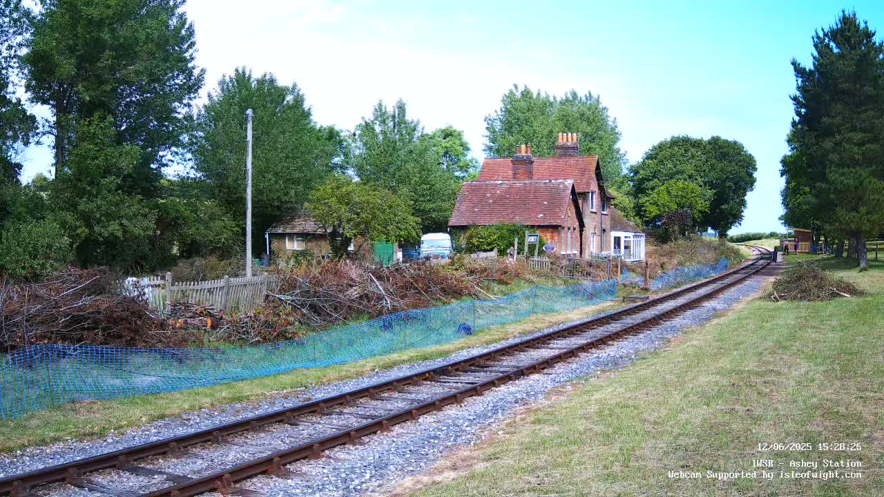 A section of railway track leads away from a small brick house, past a fence with recently cut branches behind a blue mesh barrier, on a sunny day.