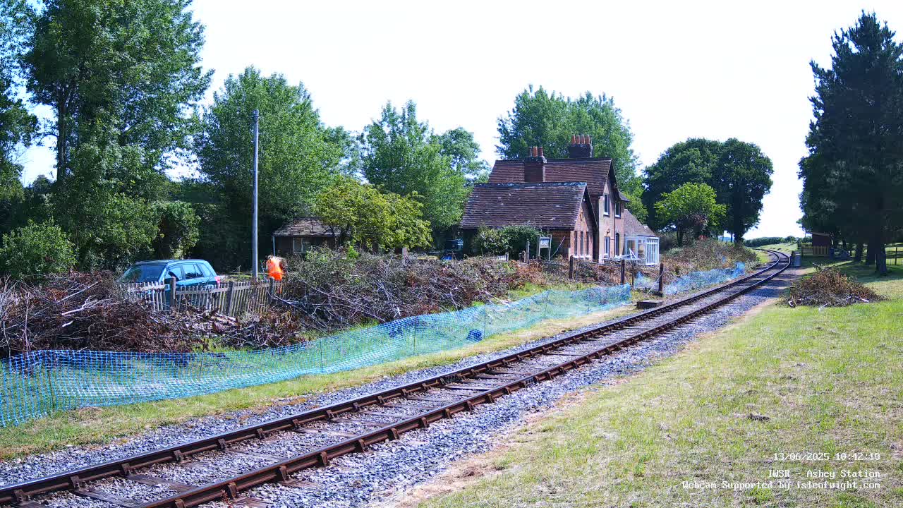 A railway line runs past a house and a pile of brush behind a blue mesh fence, on a sunny day.