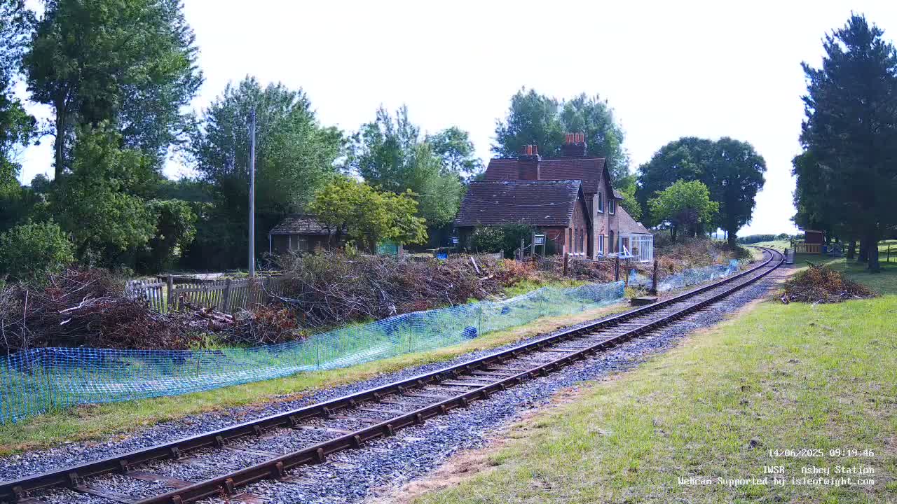 A railway track stretches into the distance past a small brick building nestled amongst trees on a partly sunny day.