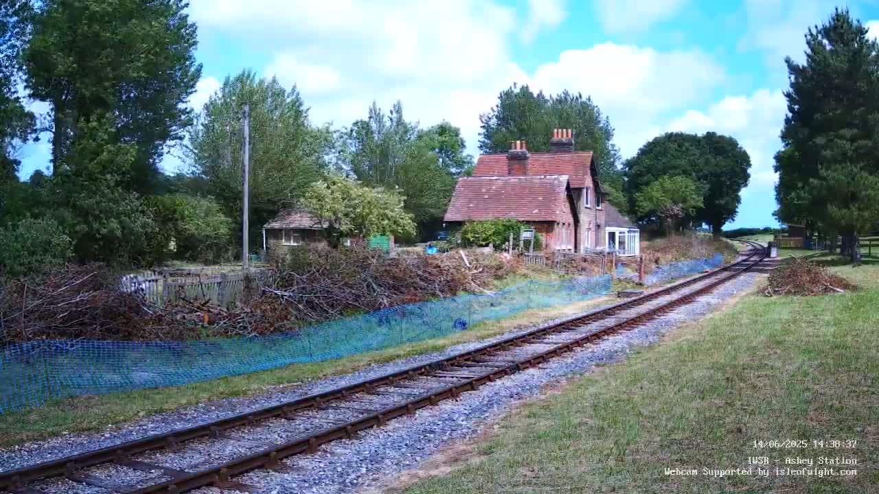 A railway track winds through a grassy area toward a brick house and other buildings nestled among lush green trees under a partly cloudy sky.