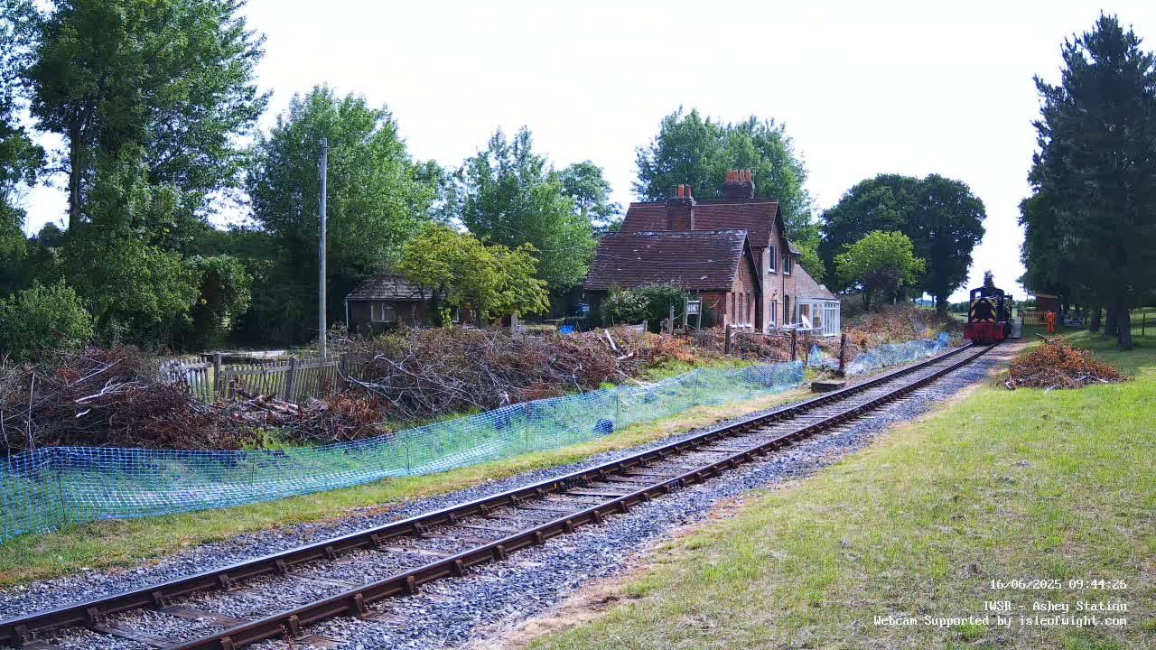 A small steam train is approaching a small brick house next to railroad tracks on a partly sunny day.