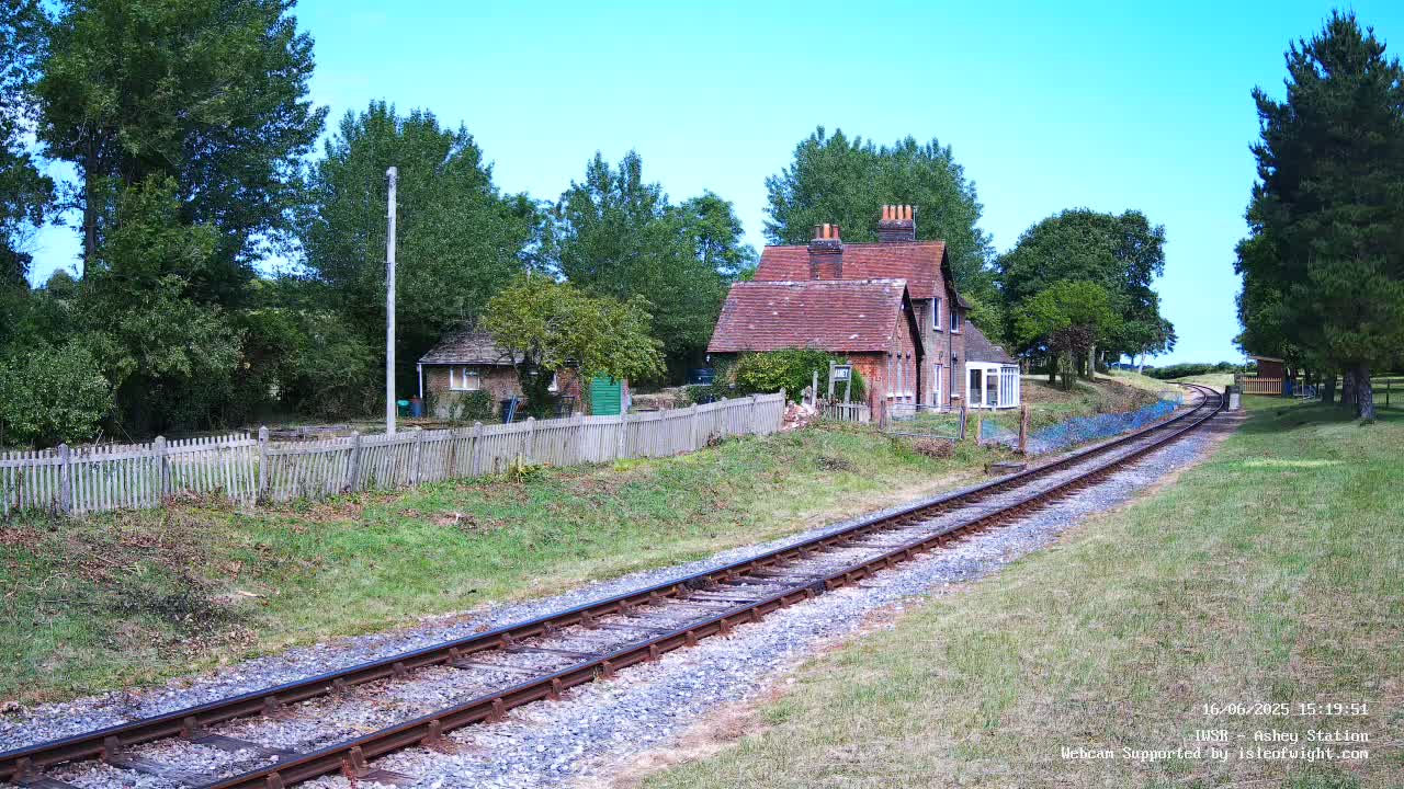 A railway track extends from a small brick house and garden, enclosed by a wooden fence, under a clear blue sky.
