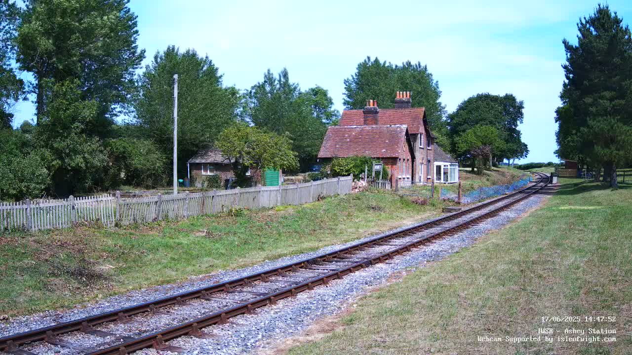 A red-roofed house sits beside a railway track on a sunny day, surrounded by green grass, trees, and a wooden fence.