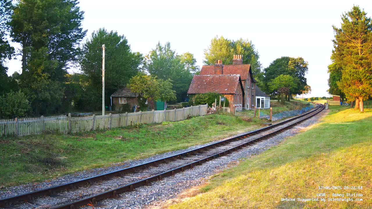 A brick house sits beside a railway line on a sunny day, surrounded by green grass and trees.