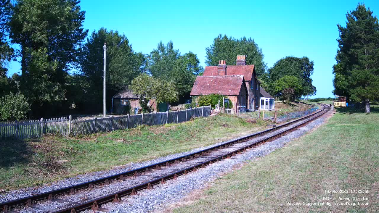 A red-roofed cottage sits beside a railway line on a sunny day, surrounded by green grass, trees, and a wooden fence.