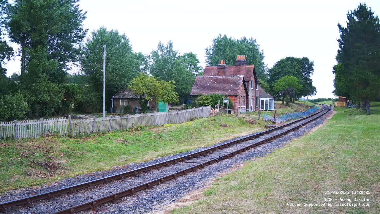A railway track extends into the distance, passing by a small brick house and outbuildings, surrounded by trees and a wooden fence under an overcast sky.