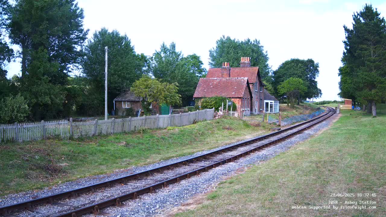 A narrow-gauge railway track leads toward a small brick house and outbuildings behind a wooden fence, under a partly cloudy sky.