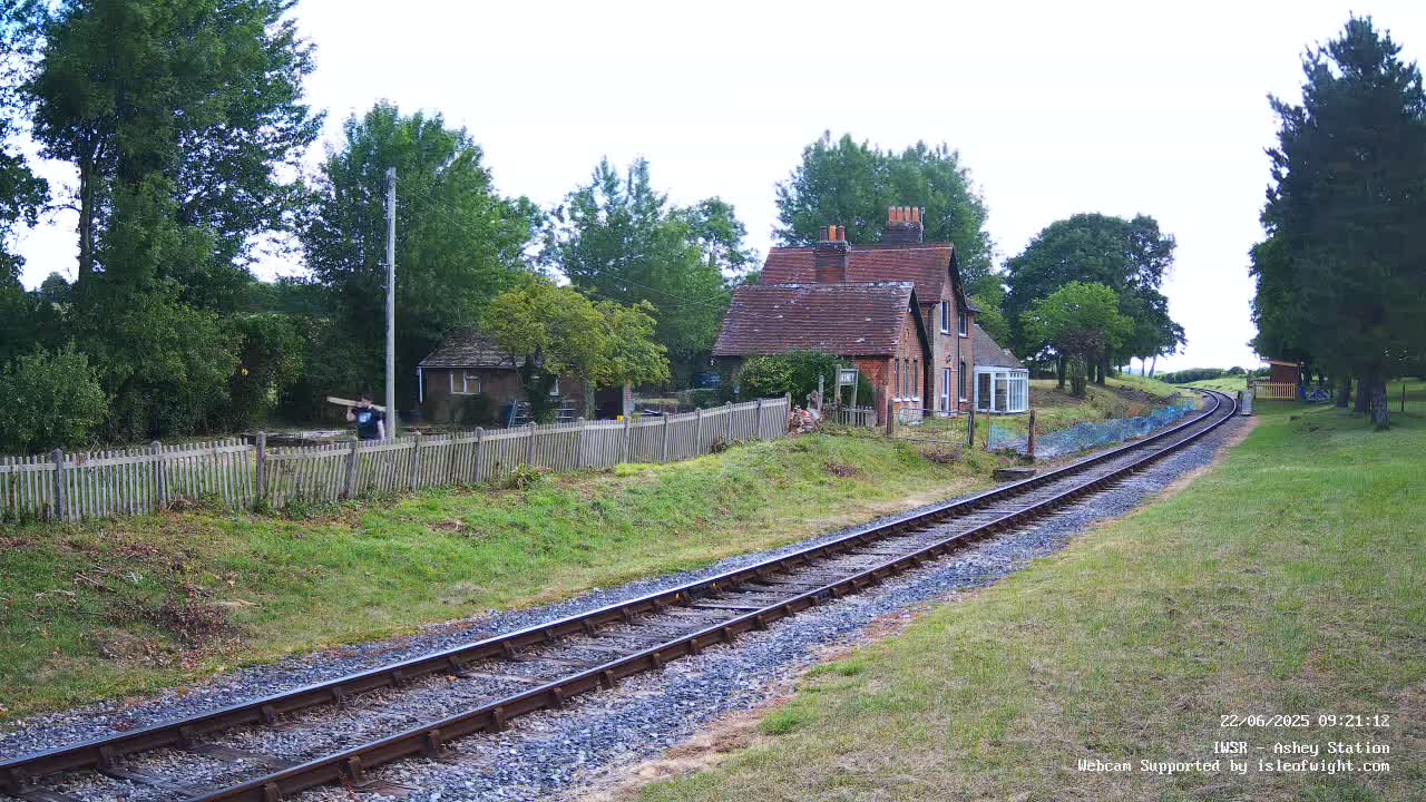 A person carries an object near a small brick house and wooden fence beside a railway track on an overcast day.
