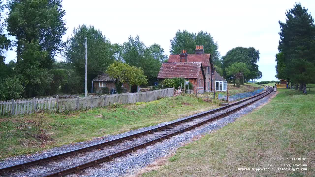 A railway track extends from a small brick building and wooden fence, passing through a grassy area lined with trees under an overcast sky.