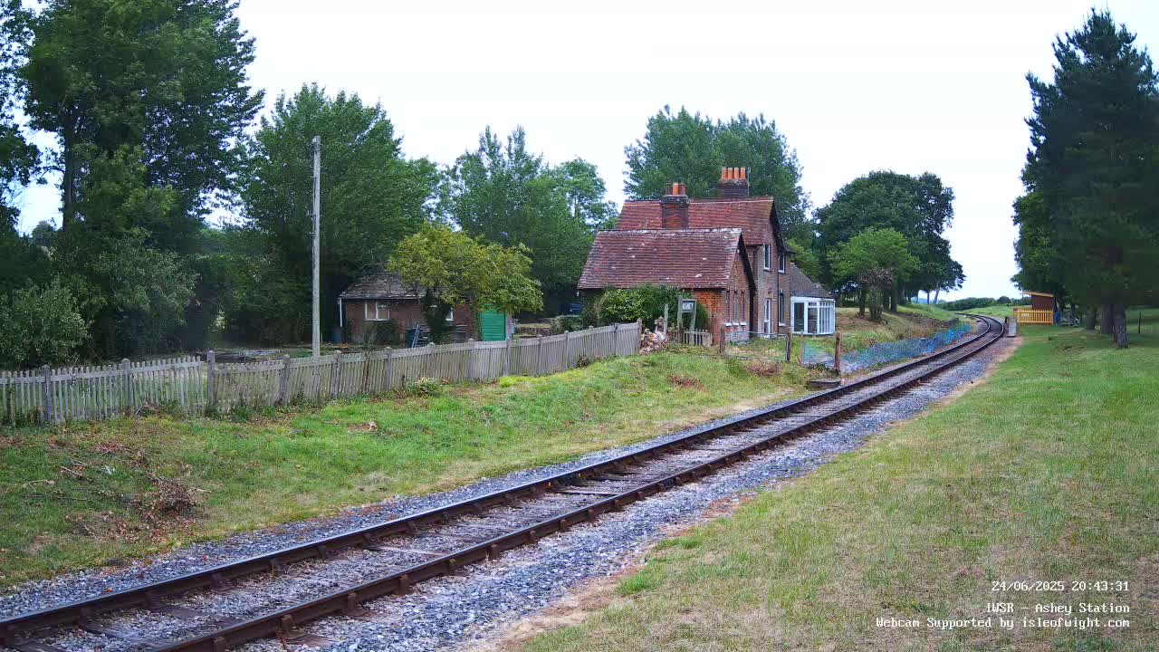 A railway track curves gently away from a small brick building and wooden fence, under an overcast sky.