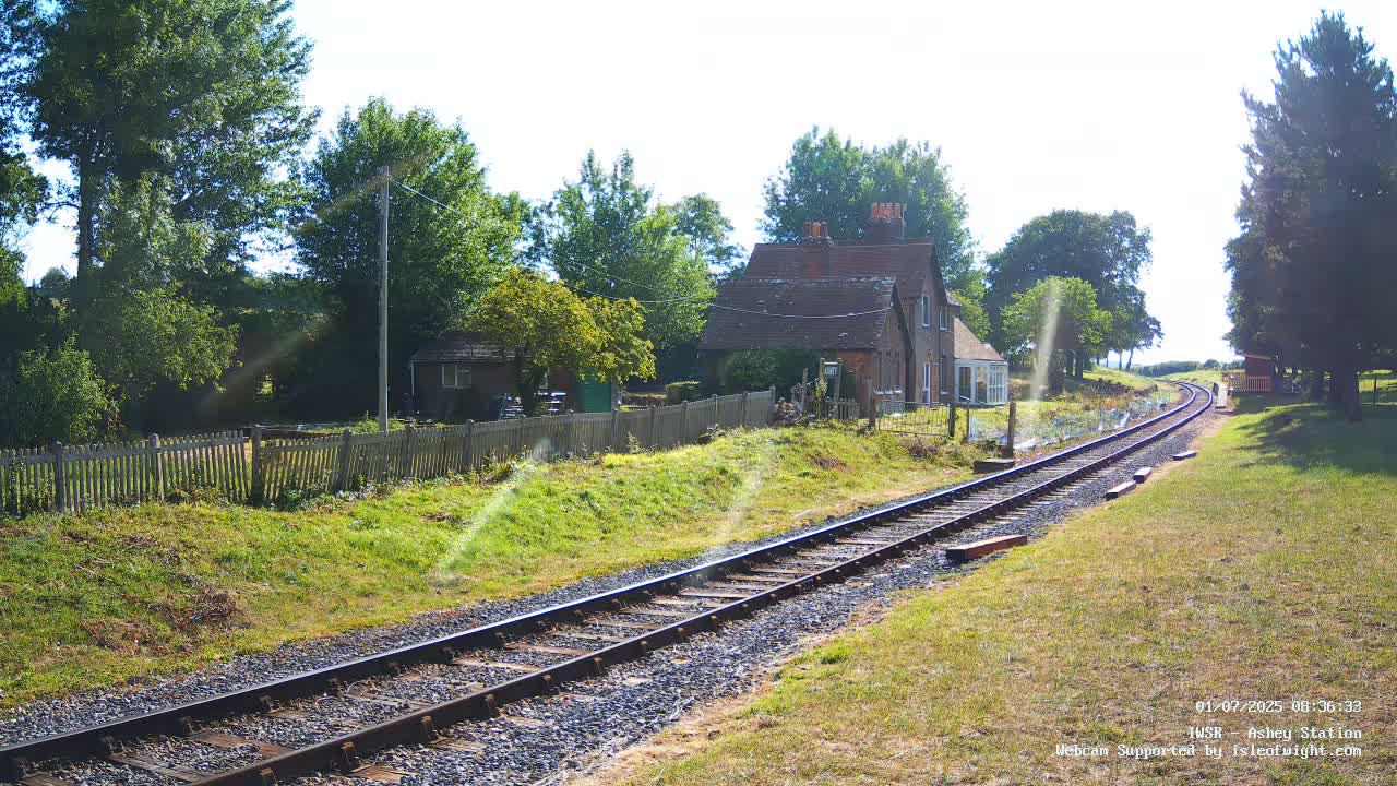 A sunny day reveals a train track leading to a small station building nestled amongst trees and a wooden fence.