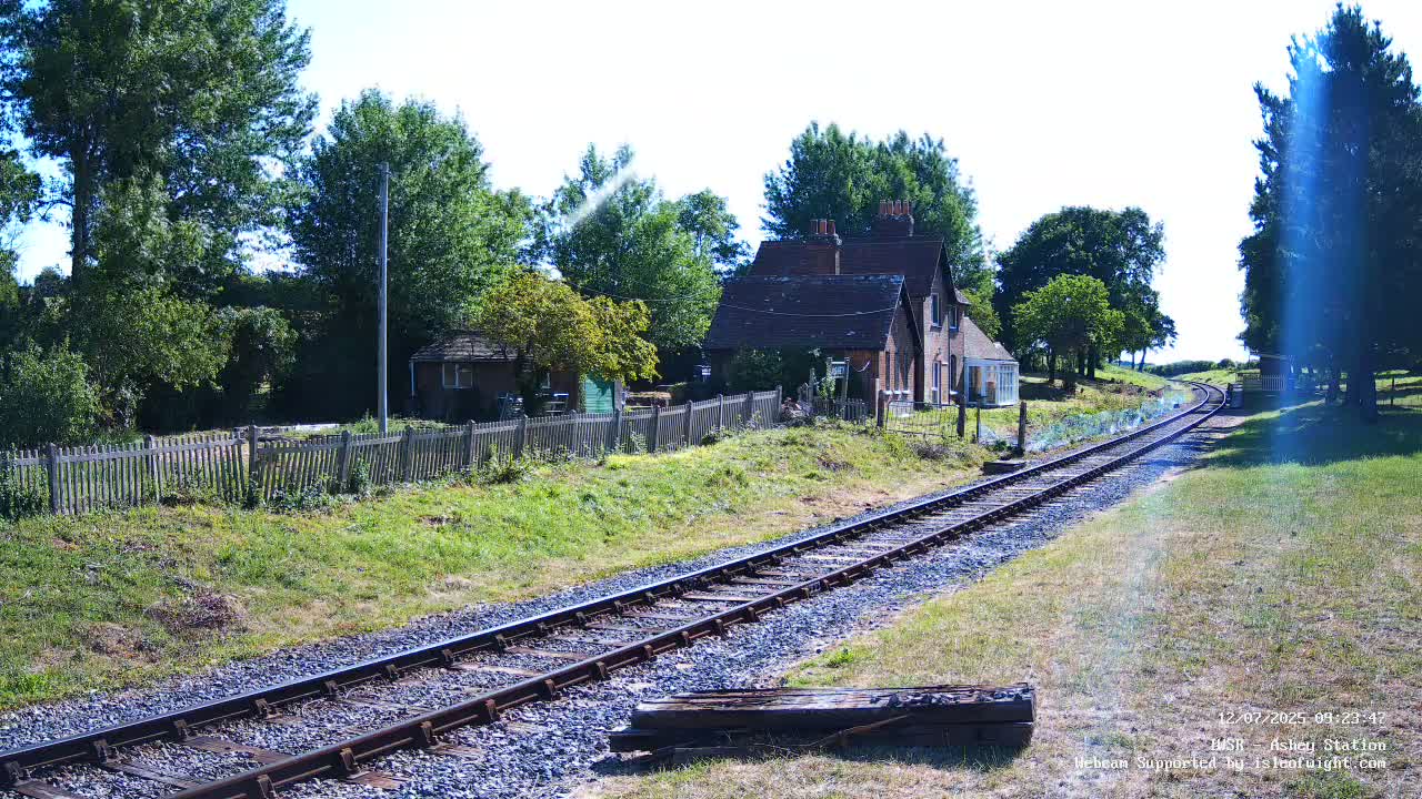 A railway track extends from a small brick building and wooden fence, into a sunny landscape dotted with lush green trees.