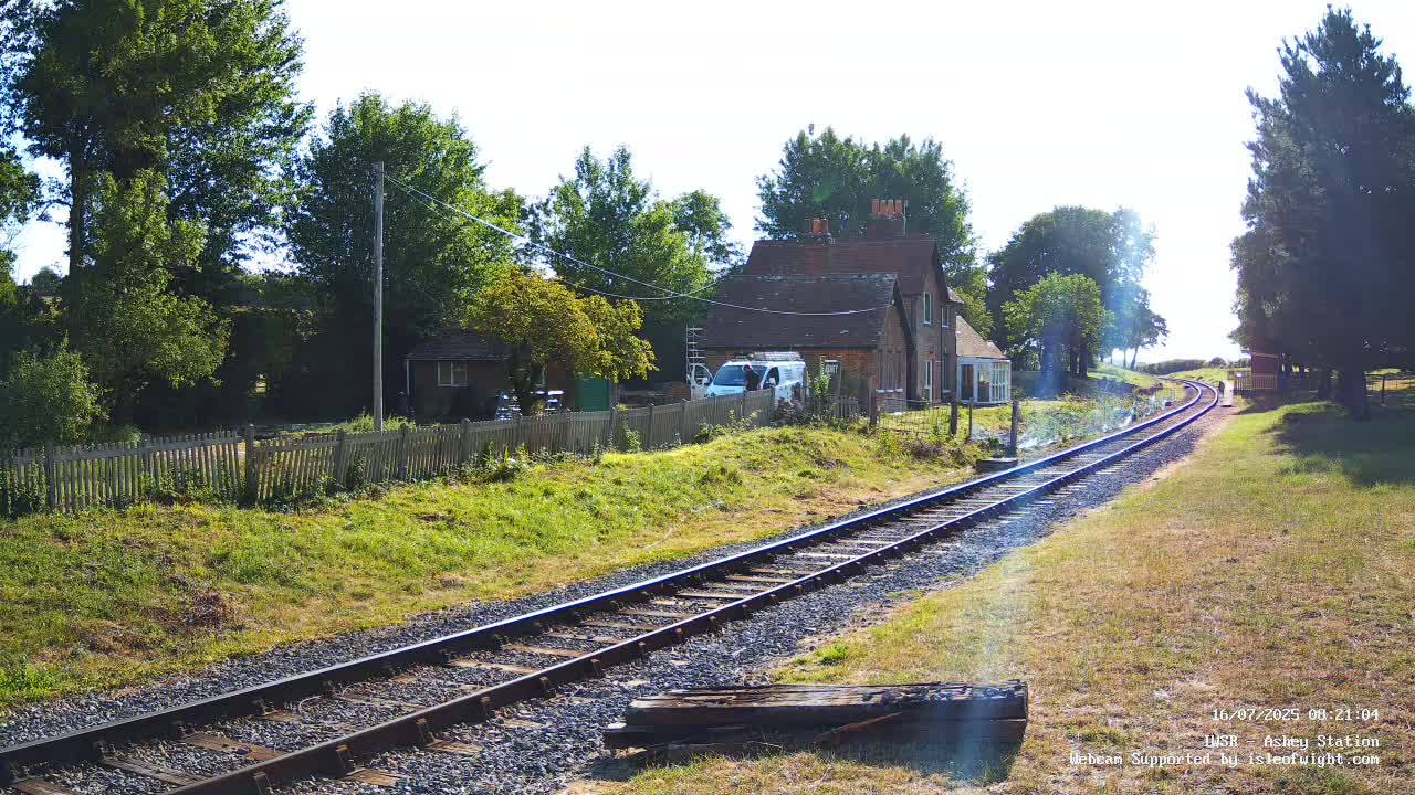 A sunny day reveals a railway track curving gently through a grassy area, past a small house and outbuildings, bordered by trees and a wooden fence.