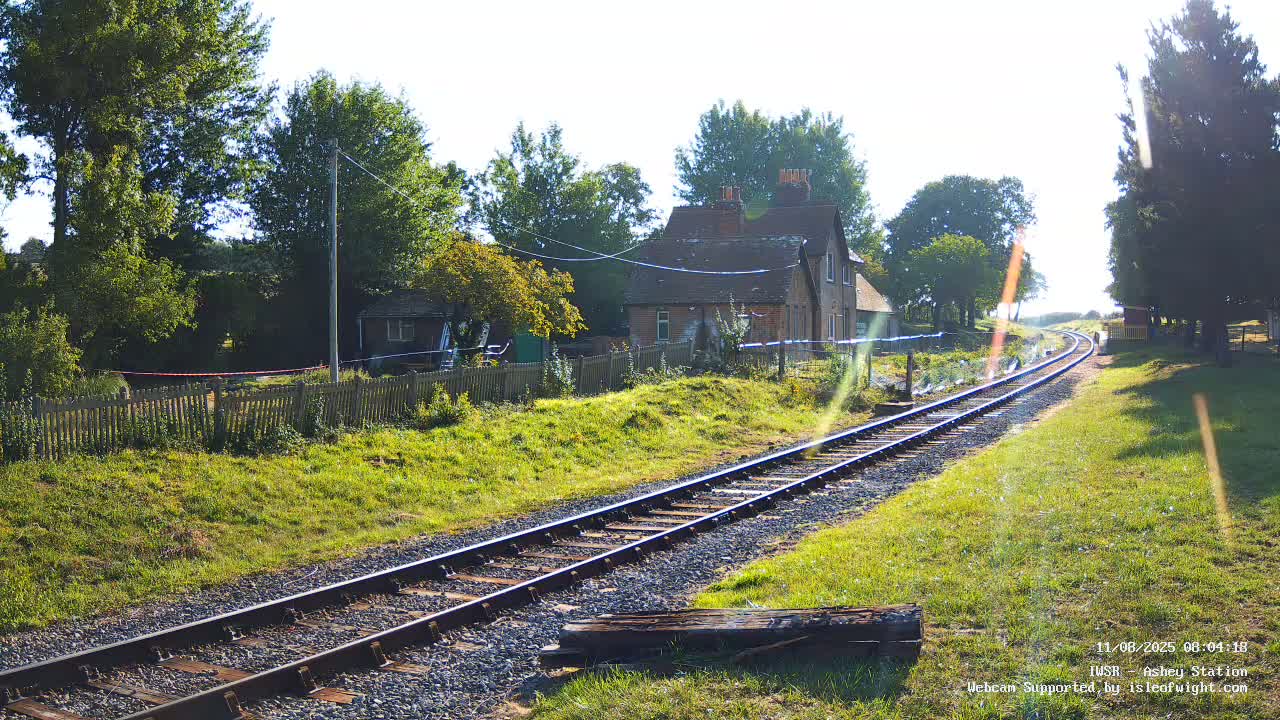 A railway track on a sunny day extends toward a small house and trees beyond a grassy embankment.