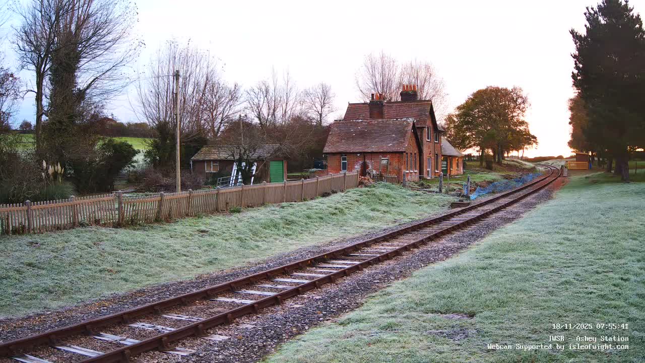 A railway track on a sunny day extends toward a small house and trees beyond a grassy embankment.