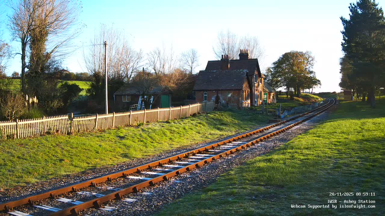 A rural railway track stretches towards a distant curve alongside a brick building and green, frost-dusted fields under a clear blue sky on a bright, cold morning.