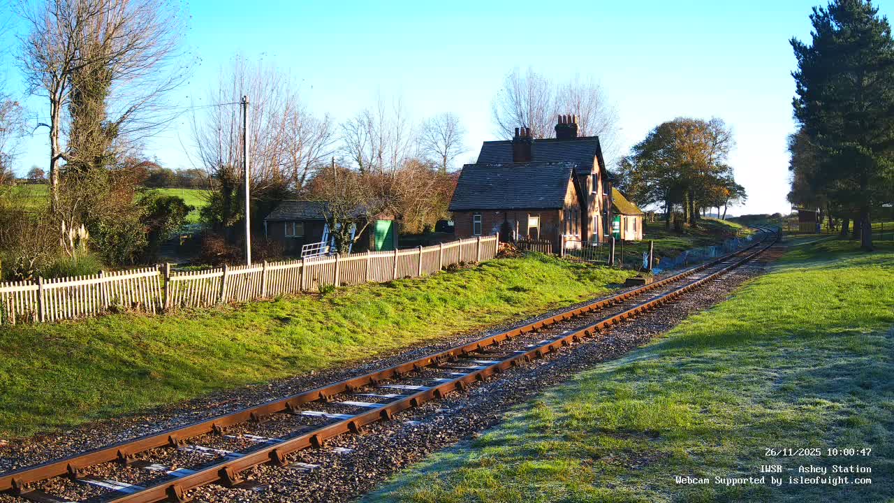 On a bright, frosty day, a railway track curves through a green, rural landscape, past a traditional brick station house, a white picket fence, and bare trees under a clear blue sky.