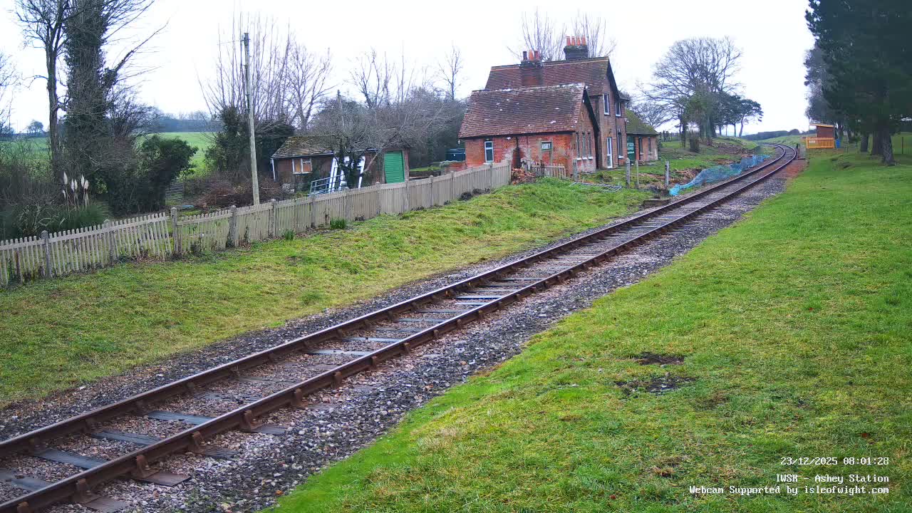 A rural railway track curves through a green, grassy landscape with a brick station house and a wooden fence on the left, under an overcast sky.