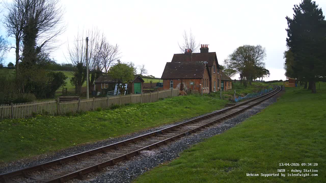 A rural railway track curves through a green, grassy landscape with a brick station house and a wooden fence on the left, under an overcast sky.