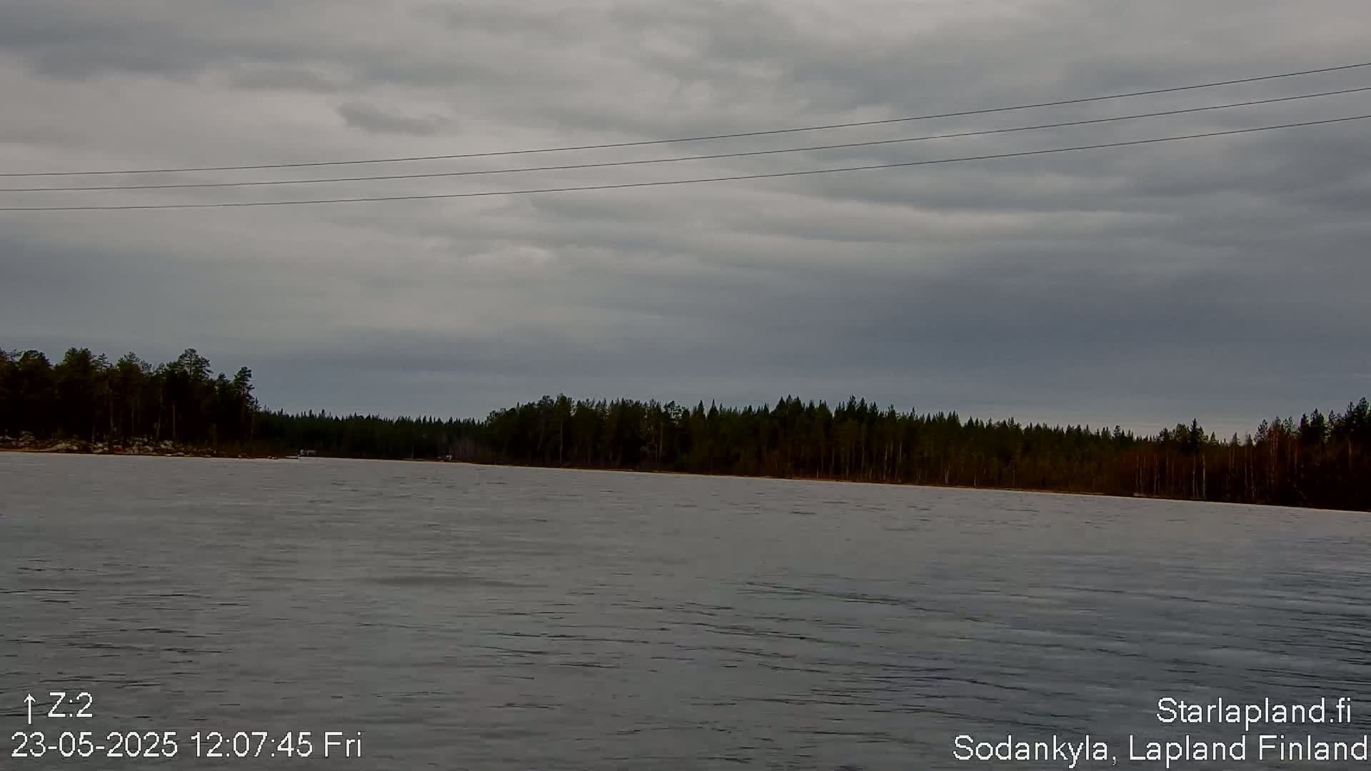 A calm body of water stretches out before a dark, overcast sky and a line of coniferous trees.