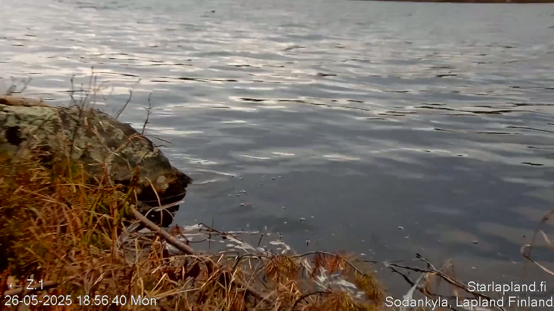 A rocky shoreline with brown vegetation borders a gently rippling body of dark water under a cloudy sky.