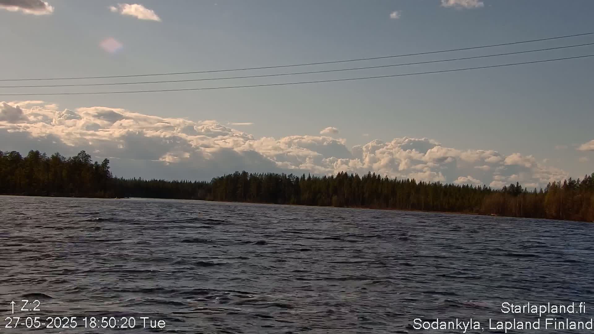 A partly cloudy sky hovers over a dark, gently rippling lake bordered by a dense evergreen forest.