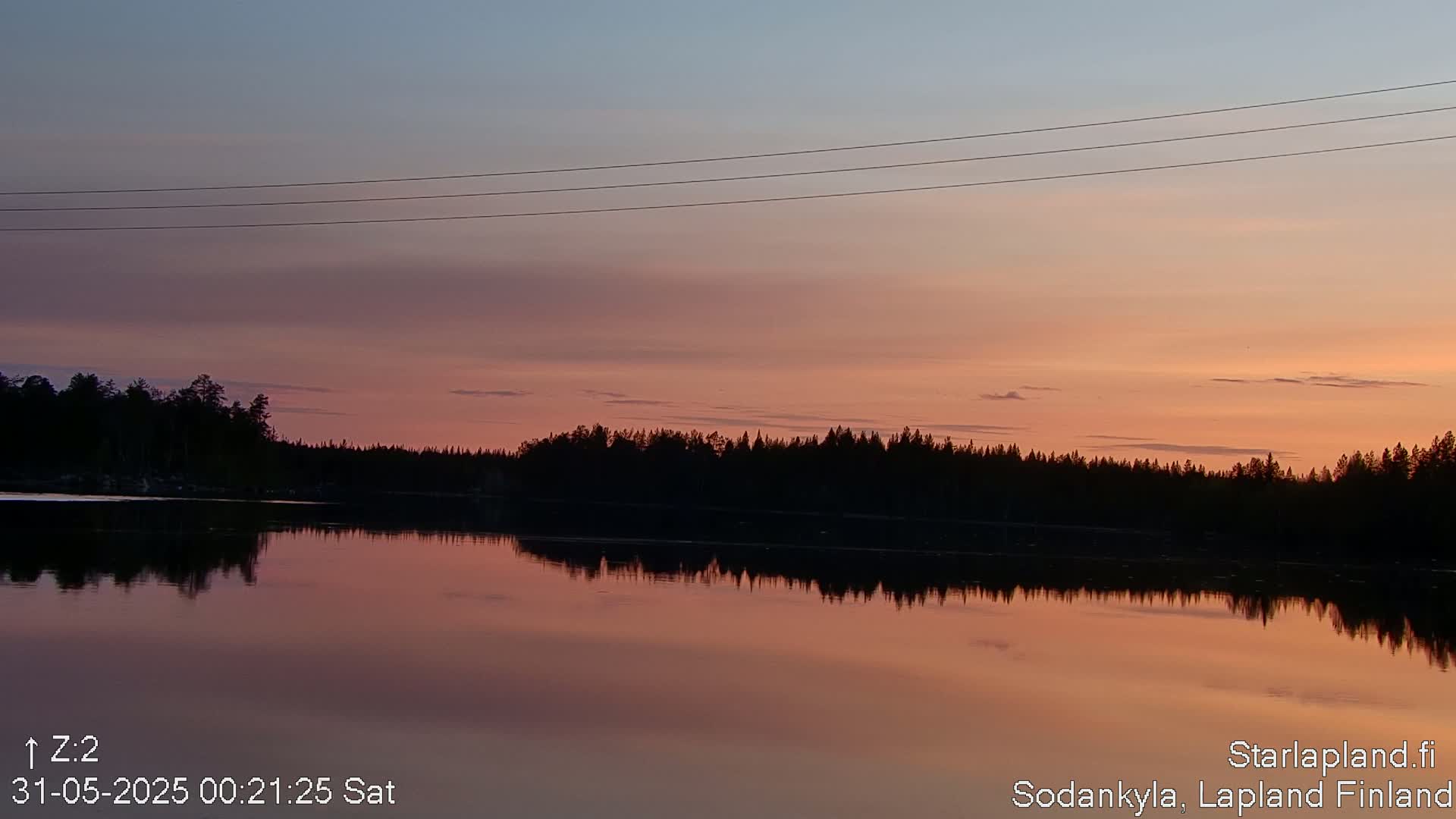 A calm lake reflects a sunset sky of soft pinks and oranges, with a dark treeline across the horizon and power lines above.