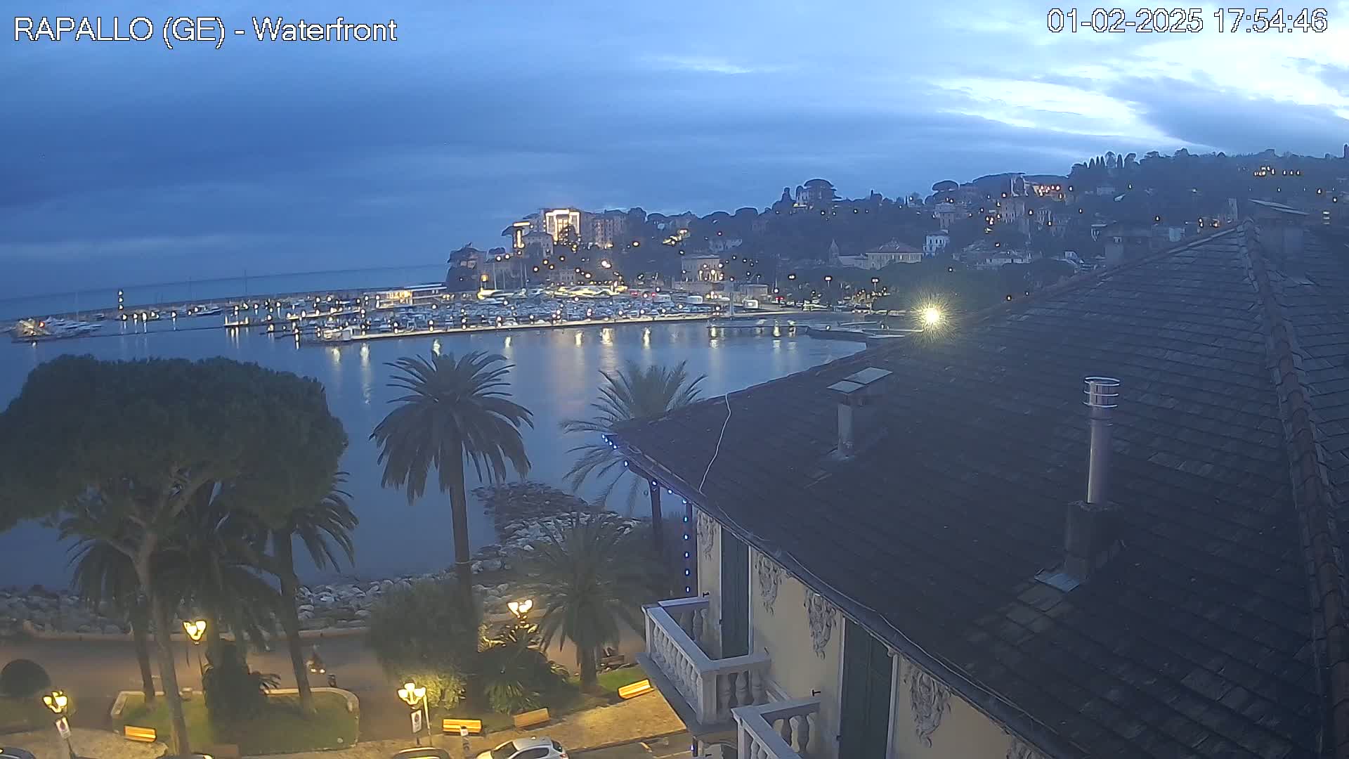 A waterfront marina filled with boats at twilight under a mostly clear, dark blue sky is viewed from above a building with a tiled roof and palm trees.