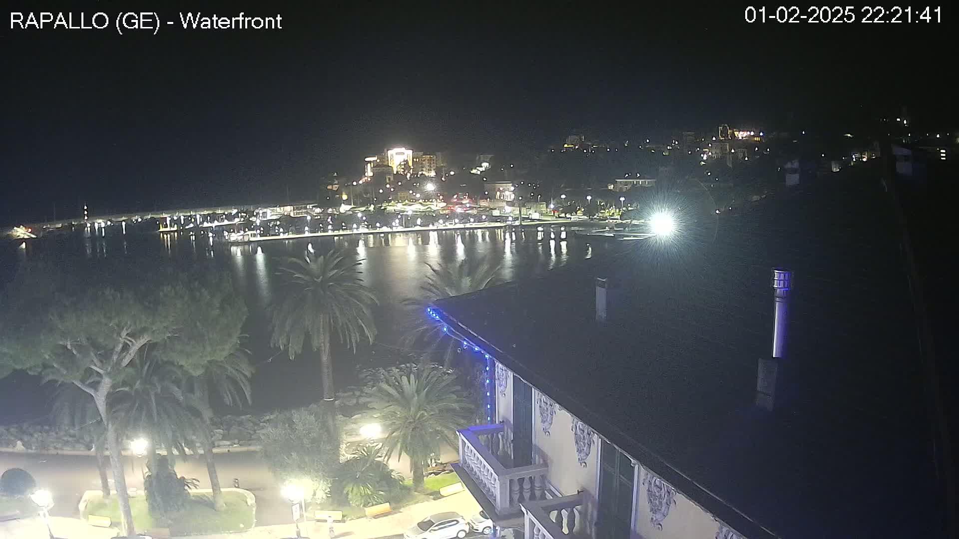 A nighttime view of a calm harbor, reflecting lights from a town on the opposite shore, seen from an elevated vantage point overlooking palm trees and a building with blue lights.