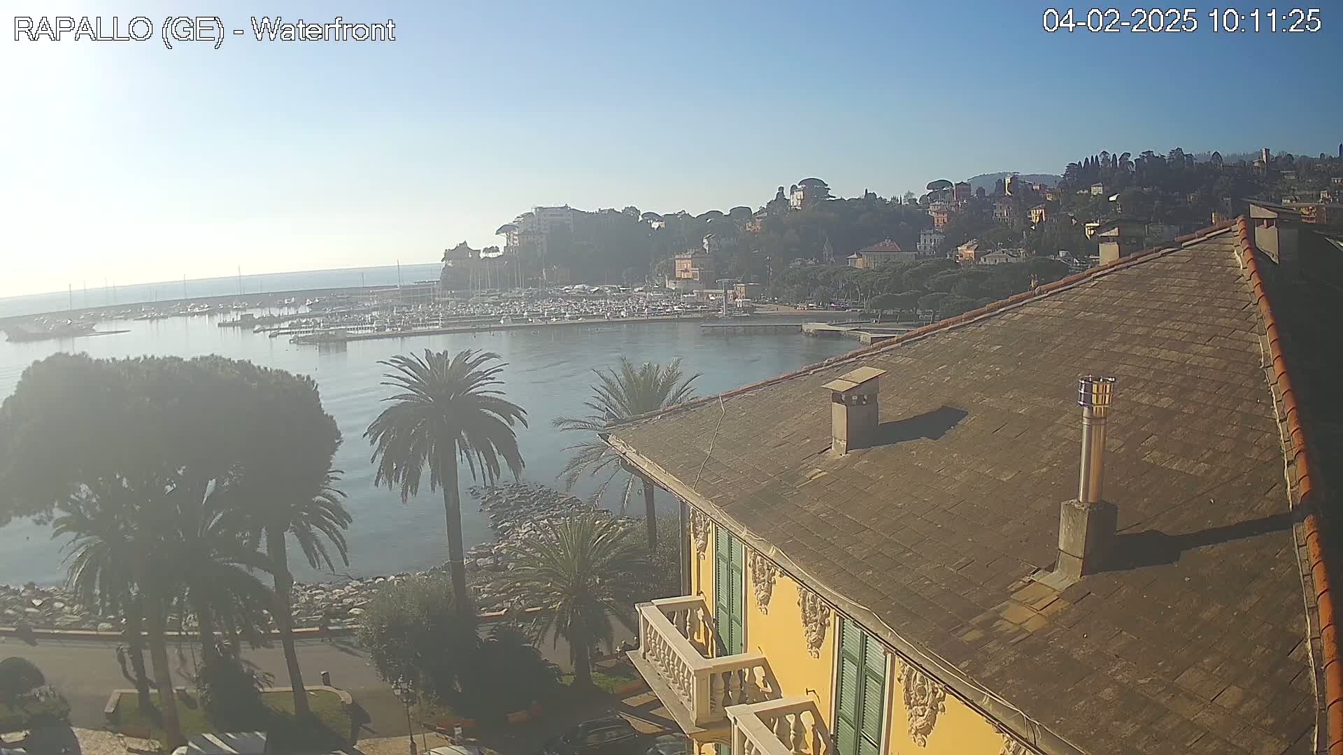 A clear, sunny day overlooks a harbor filled with boats, a waterfront town nestled on a hillside, and a portion of a yellow building's roof.