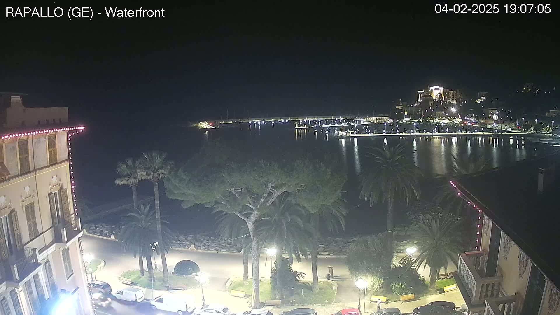 A nighttime waterfront view shows a calm body of water reflecting lights from buildings and boats in a harbor, with palm trees and buildings illuminated along a paved walkway.