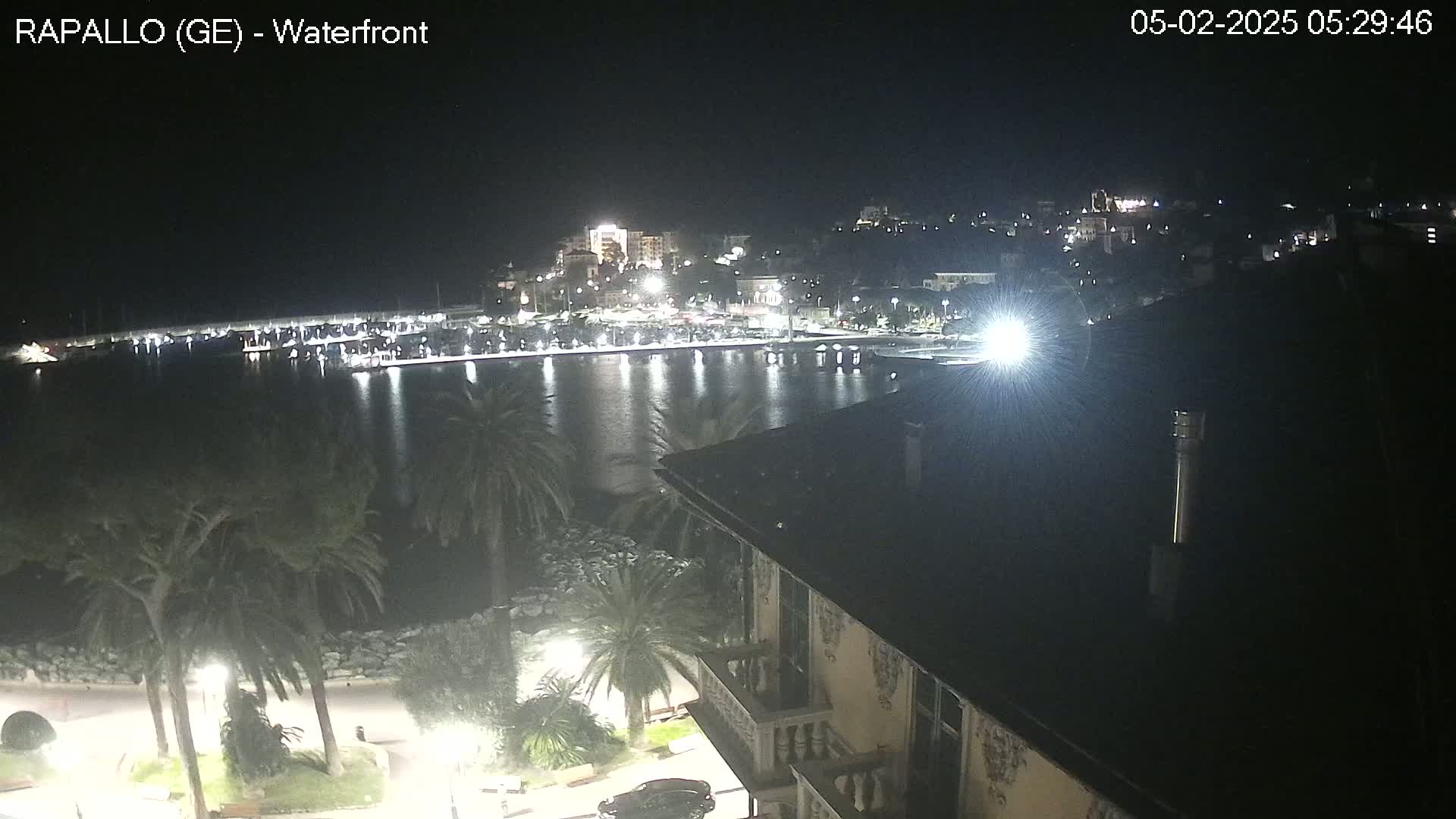 A nighttime view of a calm harbor with a town's lights reflected on the water, seen from a building overlooking palm trees and a waterfront promenade.
