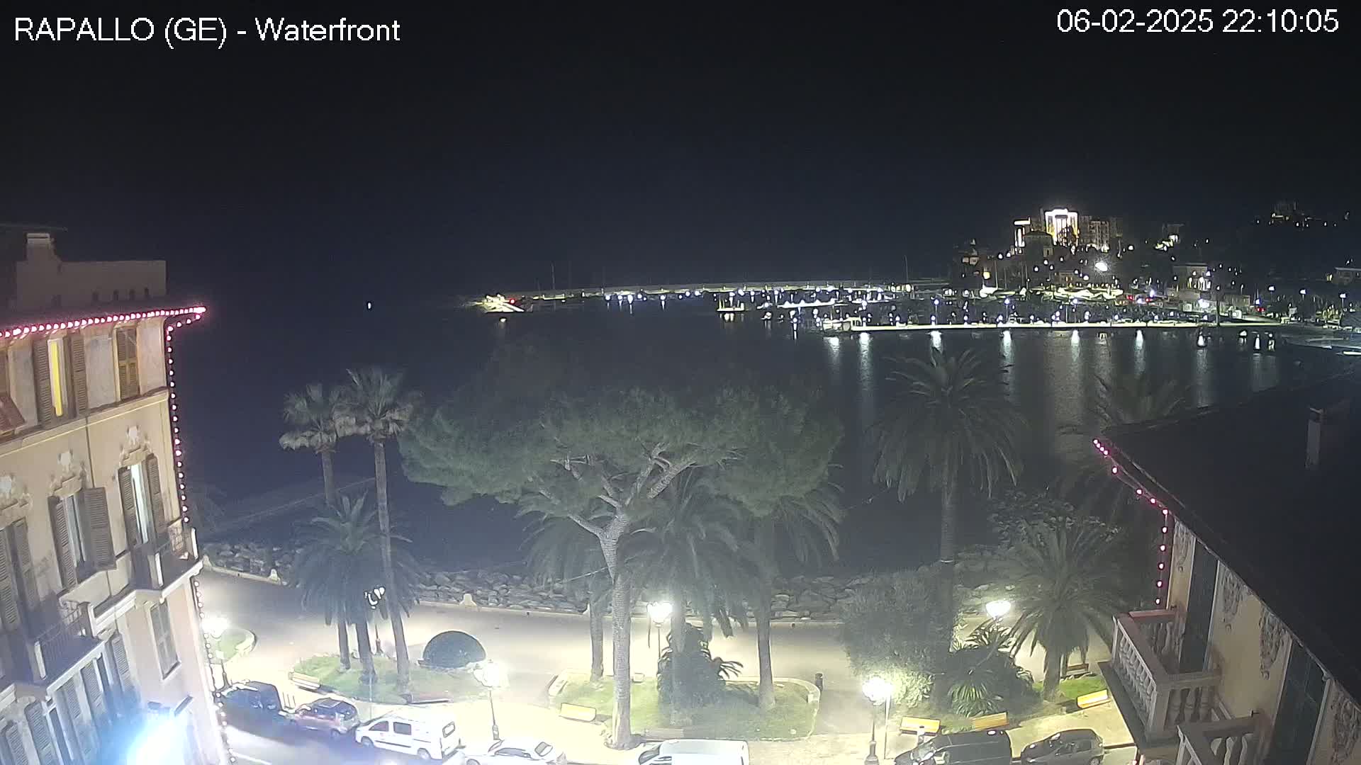 A nighttime view of a waterfront town, showing a calm harbor filled with boats, palm trees lining a paved walkway, and buildings with lights illuminating the scene.