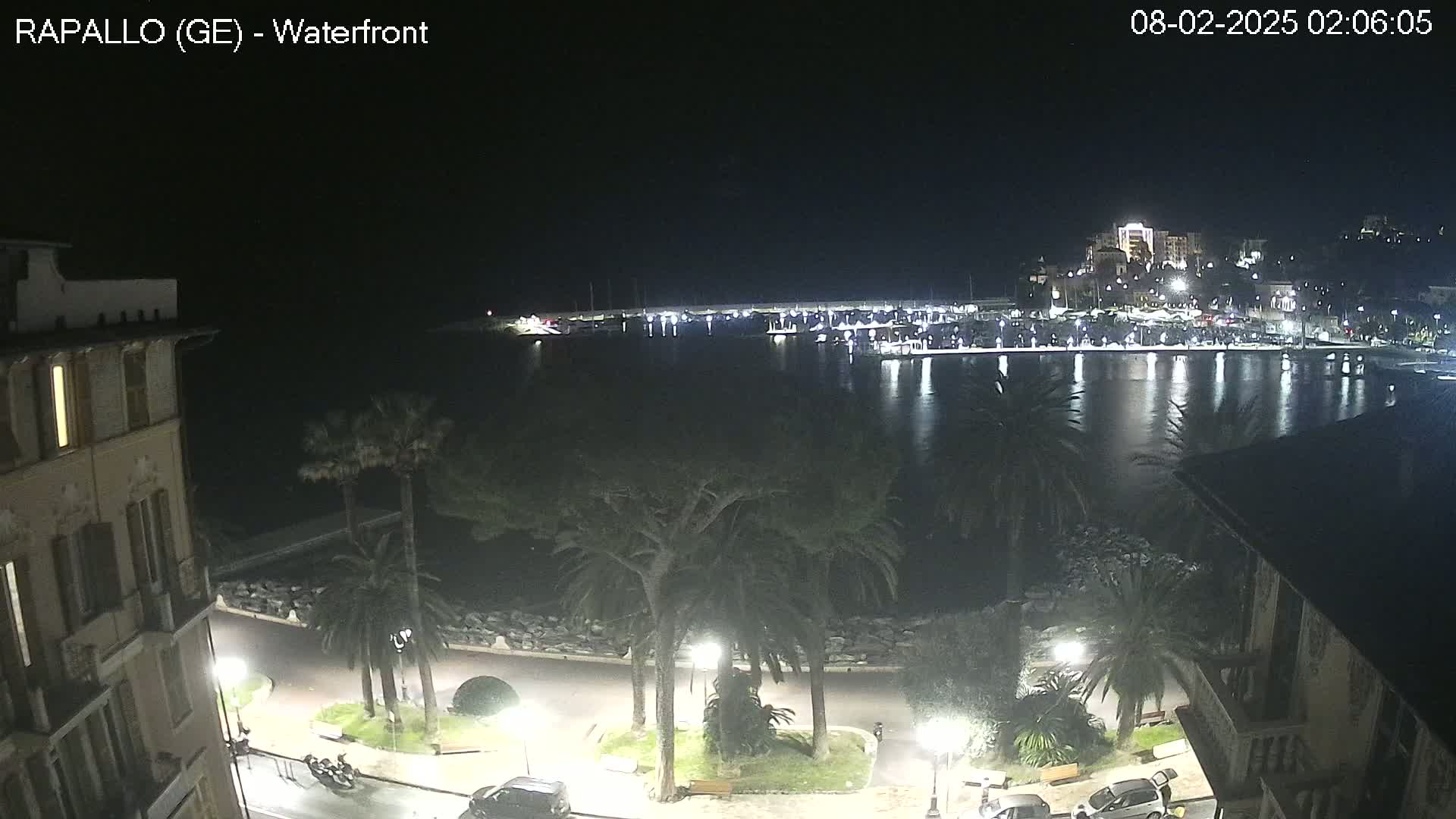 A nighttime view of a waterfront city, featuring a calm harbor filled with boats, a tree-lined promenade, and buildings illuminated by streetlights.