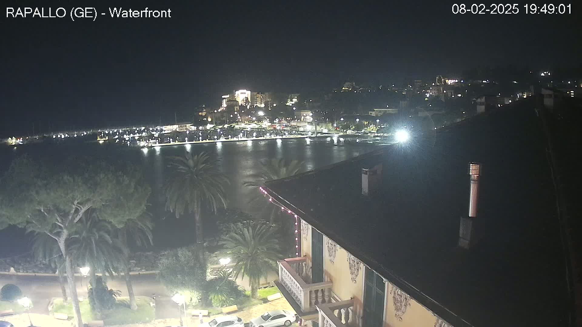 A nighttime view of a harbor filled with boats, a town lit up on a hill, palm trees, and a building with a balcony in the foreground.